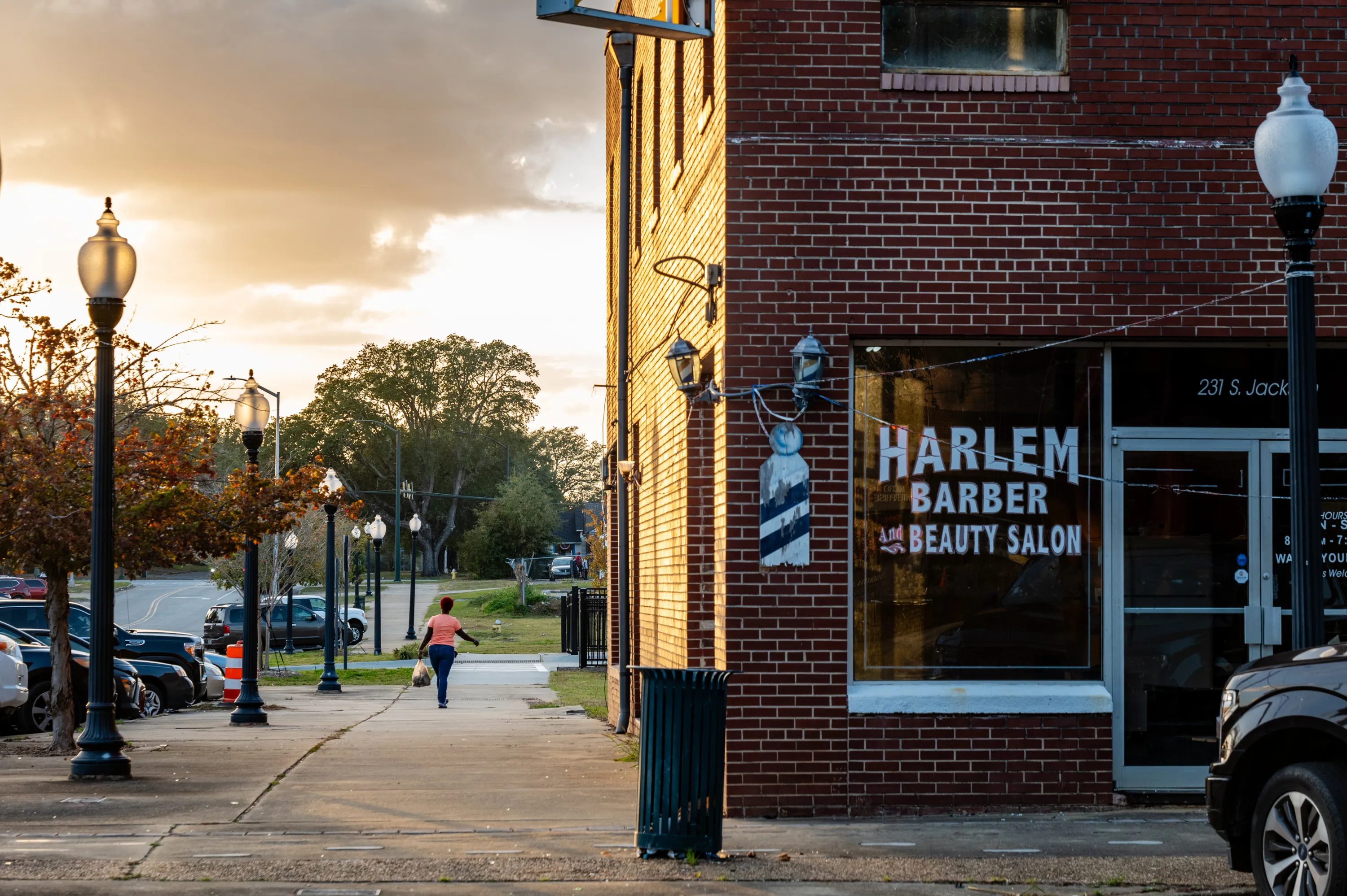 A soft light shines on the side of a building and sidewalk where a woman is walking with a grocery bag and there are cars parked in a line. The front of the building has a window labeled “Harlem Barber and Beauty Salon.”