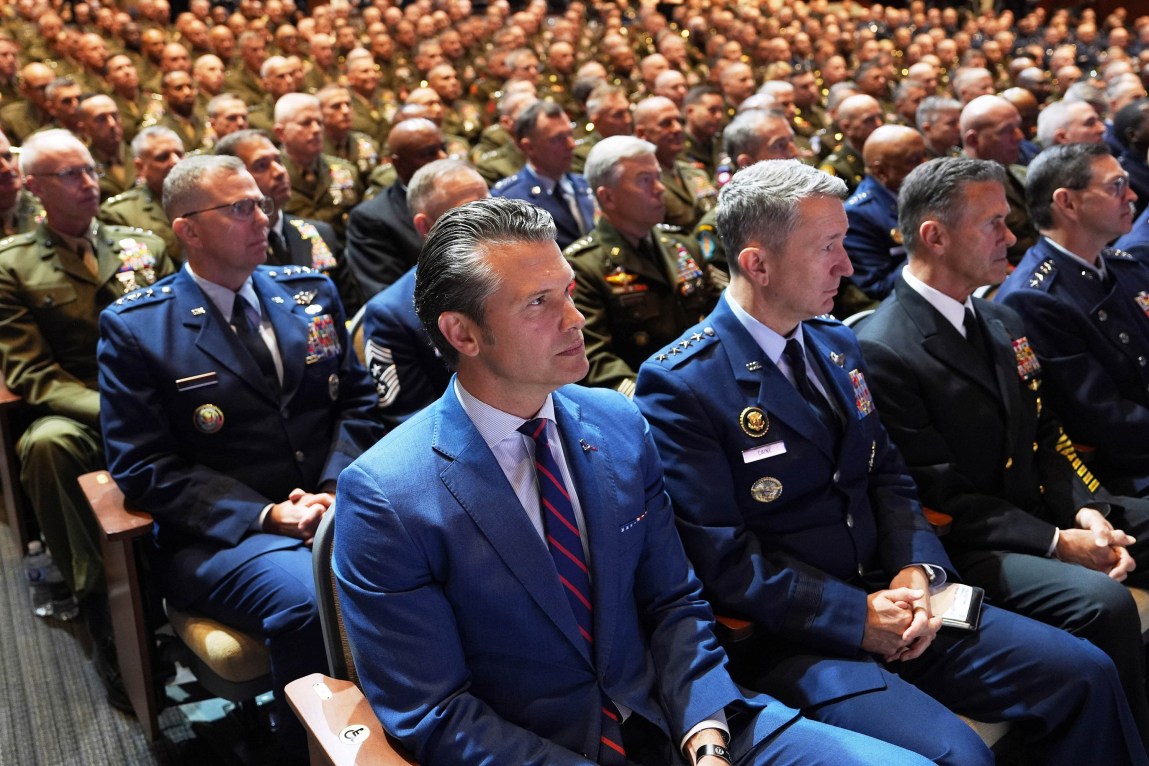 A man in a dark suit sits in the front row of an auditorium full of people in military uniforms.