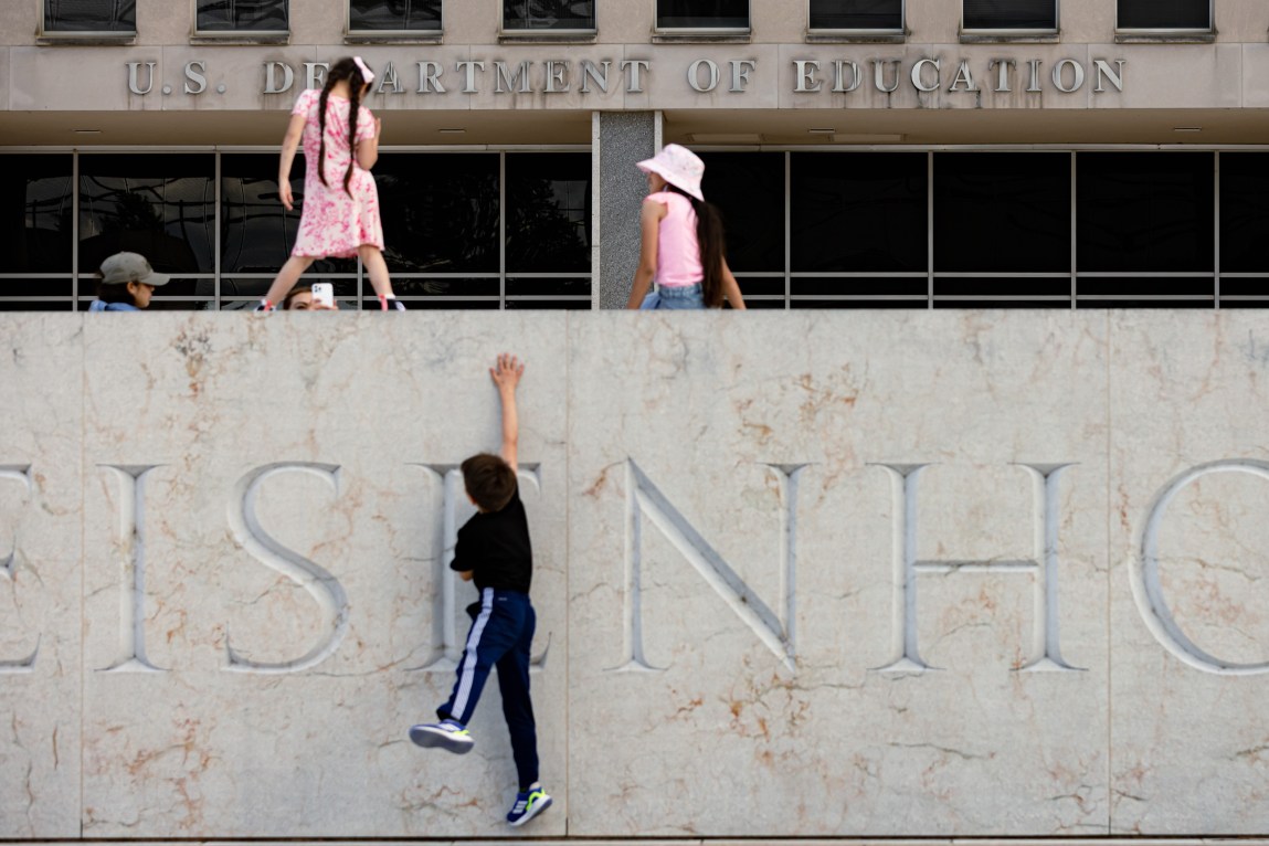 A child wearing a pink dress stands on a wall, while another child wearing a pink tank top and bucket hat sits nearby. In the background, a person holds a smartphone pointed toward them. A third child stands below, jumping with a hand against the wall. Behind all of them is a building with the words “U.S. Department of Education.”