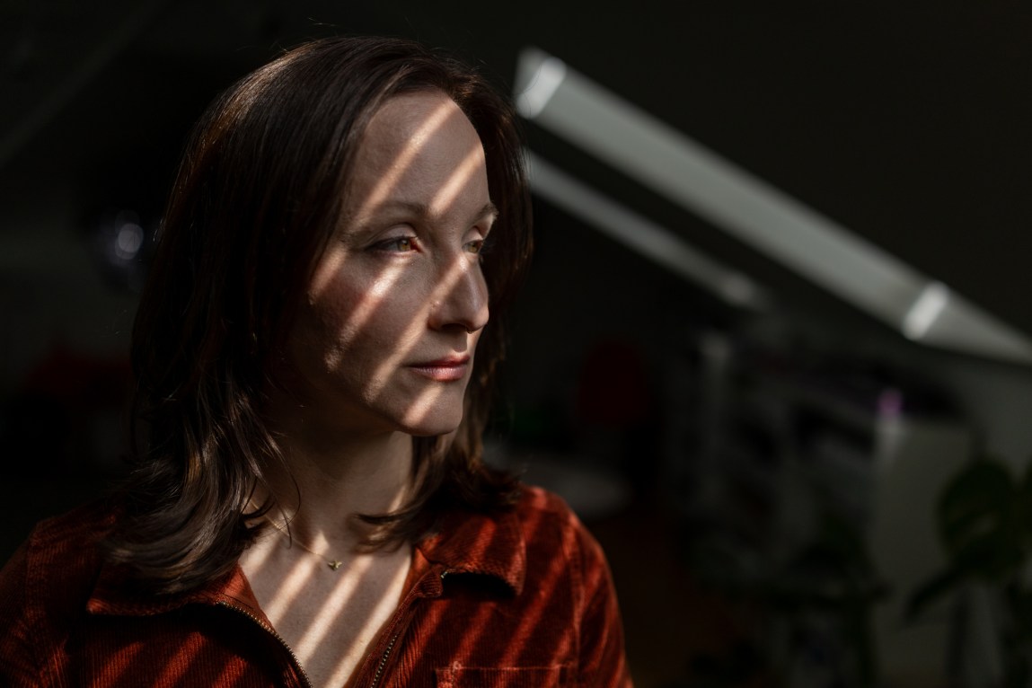 A close-up portrait of a woman looking thoughtfully to the side, with dramatic lines of sunlight and shadow from window blinds casting diagonal stripes across her face and dark orange corduroy shirt.