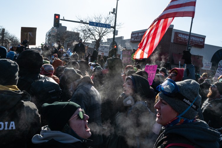 A large crowd of protesters in heavy winter clothing scream into the air at an urban intersection. Their breath steams around them.