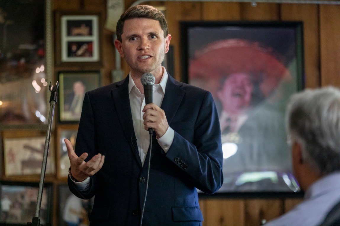 A man speaks into a handheld microphone, gesturing with his right hand as he addresses an audience inside a wood-paneled room. Framed photos and artwork decorate the wall behind him.