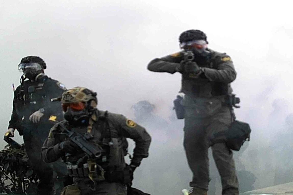 A federal agent in a uniform aims a weapon at the camera. Two other agents wearing gas masks and uniforms are to the agent’s right. There is smoke and outlines of more people behind them.