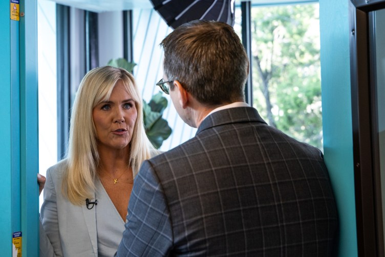 A woman with shoulder-length blond hair stands in a blue doorway speaking with a man wearing a gray checkered suit jacket.