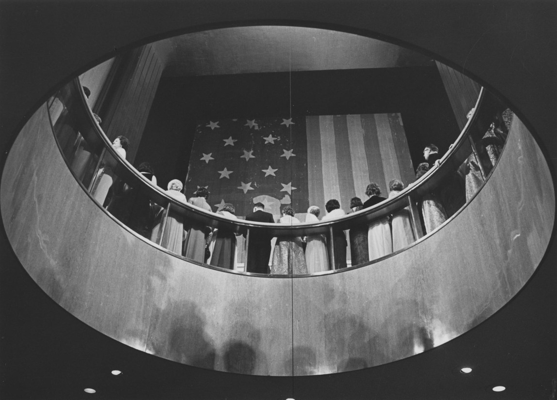 A black-and-white photo of people gathered in front of a large American flag, seen from a lower level through a circular opening.