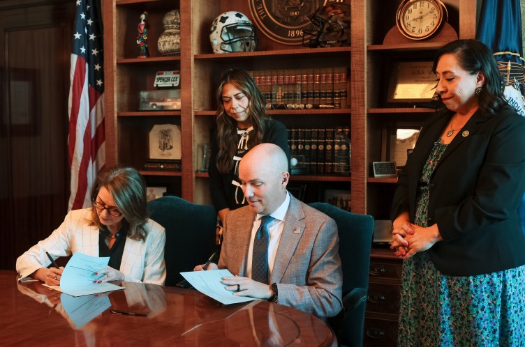 Two people, one man and one woman, sign documents at a table while other people look on, in a government office.