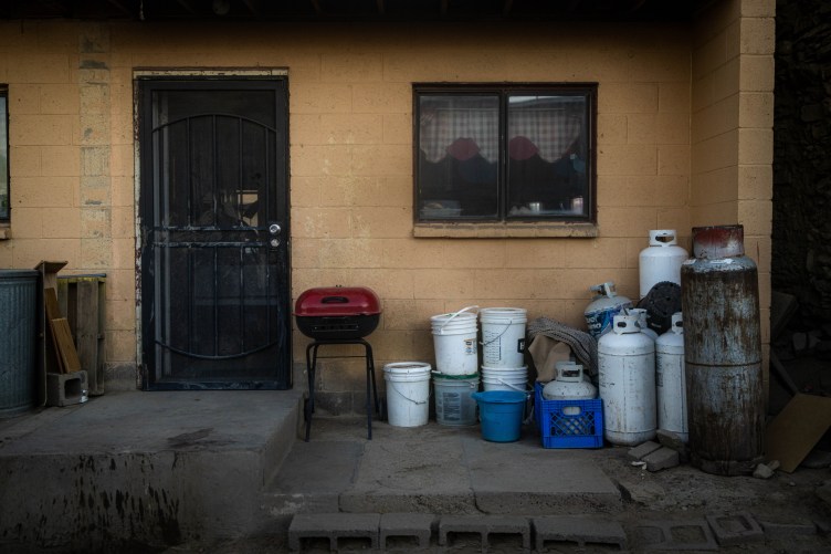 On the concrete patio of a modest home sit a collection of white and blue utility buckets, several white and rusted propane tanks and a small, red grill.