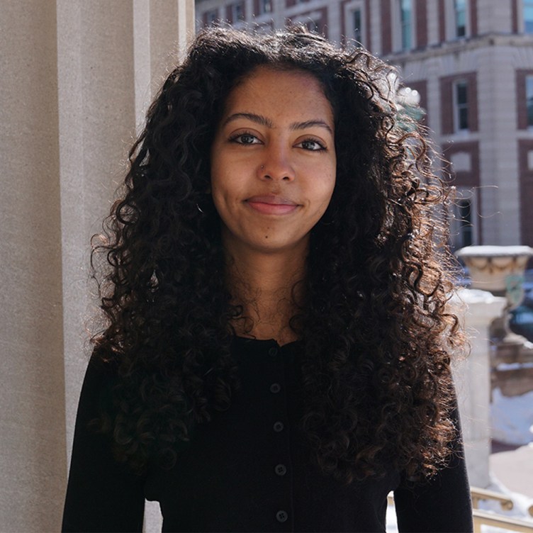 A woman with long, dark, curly hair wears a black long-sleeved shirt and smiles at the camera. She is standing in front of a white pillar, and a stone-and-brick building can be seen in the background.